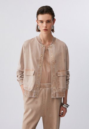 Donna con capelli corti pettinati all'indietro indossa una giacca beige con bottoni, pantaloni abbinati e grandi orecchini e braccialetti d'argento su sfondo neutro.