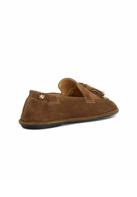 Bruine suede loafers met kwastjes, platte zwarte zool en een klein metalen logodetail aan de zijkant, weergegeven vanuit een zijaanzicht.