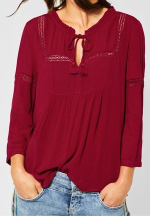 Bordeauxrode blouse met een ronde hals, sluiting met strik en kwastjes. Voorzien van kanten details en plooien, gecombineerd met lichtblauwe denim shorts.