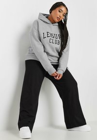 Sudadera gris con el texto "LEISURE CLUB", de corte holgado y con un bolsillo tipo canguro. Combinada con pantalones amplios negros y zapatillas blancas.