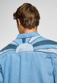 Ljusblå jeansjacka med en solmotif broderad på baksidan, med mörkblå akcenter och en liten "Lee"-logotyp.