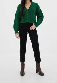 Zielony dziergany kardigan z dekoltem w kształcie litery V, z przodu zapinany na guziki i z rękawami balonowymi. Połączony z czarnymi jeansami z wysokim stanem oraz brązowymi botkami na obcasie.