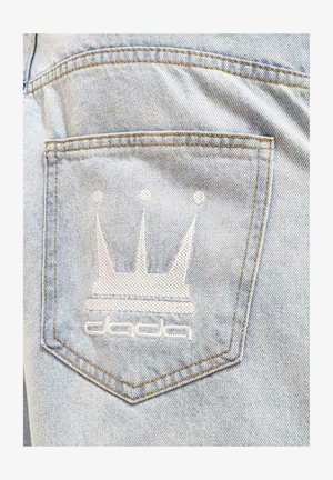 Light blue denim pocket with white embroidered crown logo and "dada" text. Features visible stitching and a textured fabric surface.