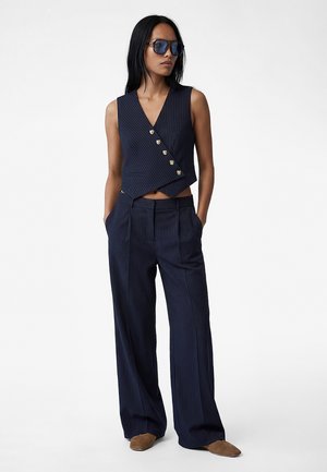 Kvinde iført navyblå ærmeløs vest med nålestriber og guldknapper samt matchende bukser med vide ben, brune sko og blå solbriller, stående med hænderne i lommerne.