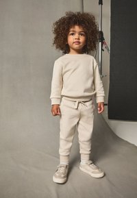 Beige collegepuku, jossa pehmeä kangas, pyöreä pääntie, resorit hihoissa, joustava vyötärö ja beiget lenkkarit harmailla yksityiskohdilla.