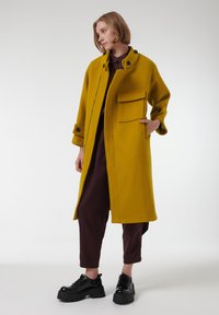 Abrigo de lana mostaza con cuello alto, botones negros y un bolsillo delantero, combinado con pantalones marrones y zapatos negros gruesos.