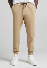 Beige Jogginghosen aus glattem Material, mit elastischem Bund und Kordelzug, schmal zulaufenden Knöcheln und seitlichen Taschen. Kombiniert mit weißen Sneakers.