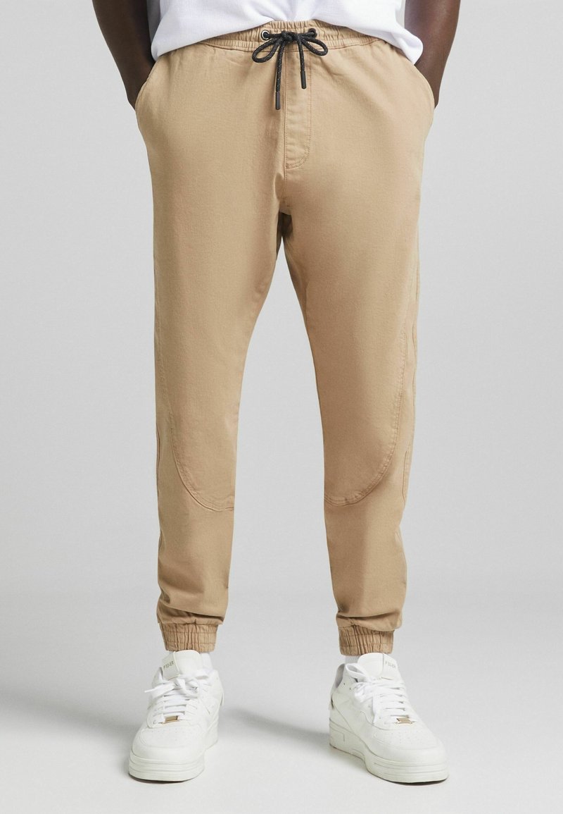 Beige Jogginghosen aus glattem Material, mit elastischem Bund und Kordelzug, schmal zulaufenden Knöcheln und seitlichen Taschen. Kombiniert mit weißen Sneakers.