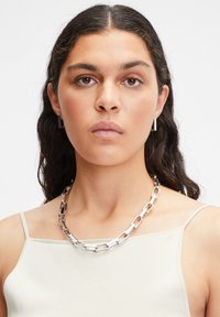 Sølvmetal kædehalskæde med rektangulære led, parret med enkle rektangulære øreringe, båret over en lysefarvet tanktop.