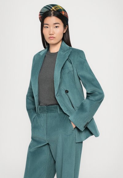 Woman in teal corduroy suit with hands in pockets, wearing a dark gray shirt and a plaid beret, standing against a white background.
