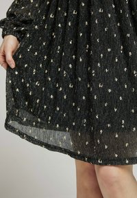 Robe noire avec éclat métallique, tissu texturé et design en étages. Présente de petits motifs floraux dorés sur l'ensemble. L'ourlet est transparent.