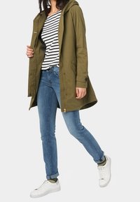 Olivově zelená kapucí opatřená bunda s knoflíkovým zapínáním, doplněná proužkovaným tričkem a modrými džínami, dokončená bílými teniskami. Casual střih.