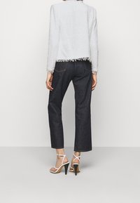 Biała teksturowana kurtka z postrzępionymi brzegami, zestawiona z ciemnymi jeansami i białymi sandałami na wysokim obcasie, widziana od tyłu.