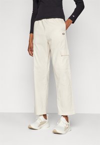 Béžové cargo kalhoty s kapsami, elastickým pasem a volným střihem, spárované s bílými teniskami. Vrchní část je černá, dlouhý rukáv.