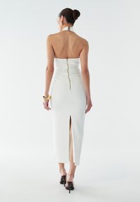 Robe blanche sans manches avec une fermeture éclair au dos et une fente au niveau de l'ourlet. Le tissu a une texture lisse ; présente un design de col halter contrastant.