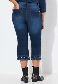 Pantalones capri de mezclilla azul oscuro con dobladillo festoneado y acentos de cuentas blancas; bolsillos traseros visibles; combinados con botas de tobillo negras con tachuelas.