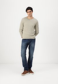 Camiseta de manga larga de canalé gris claro, vaqueros azules y zapatillas marrones. Hombre de pie con las manos en los bolsillos, mirando ligeramente hacia un lado.