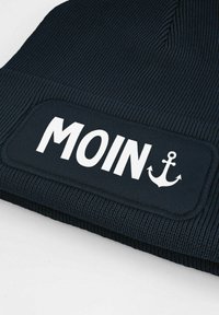 Navy beanie with a ribbed texture; features a white patch with the word "MOIN" and an anchor graphic in the lower corner.