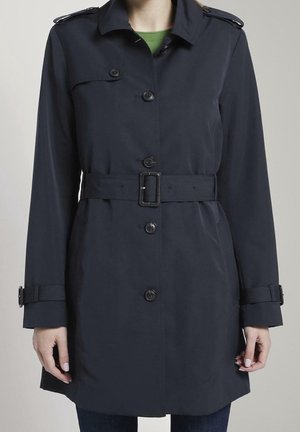 Frau trägt einen dunkelblauen Trenchcoat mit Gürtel, Knopfleiste, Schulterklappen und Manschettenriemen über einem grünen Oberteil und dunkler Hose.