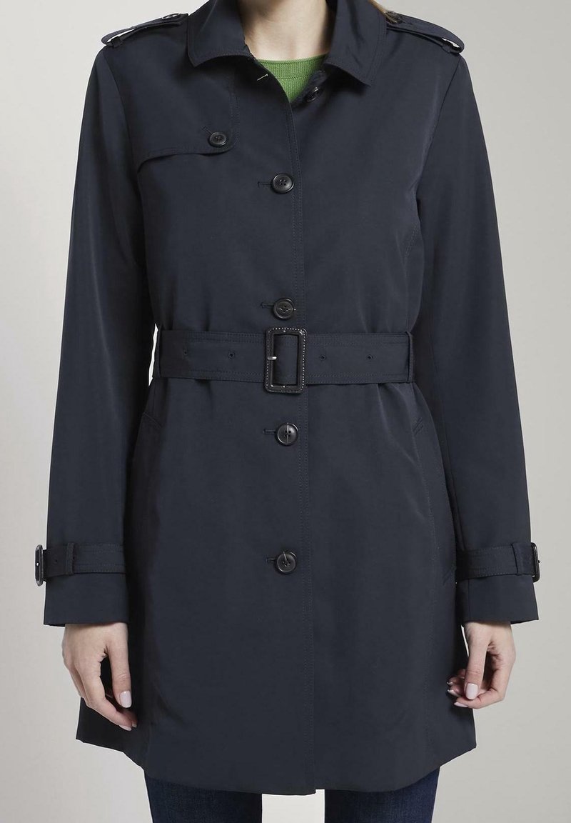 Femme portant un trench-coat ceinturé bleu marine foncé avec des boutons sur le devant, des épaulettes et des sangles aux poignets, par-dessus un haut vert et un pantalon foncé.