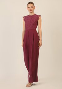Salopette bordeaux con un corpetto in pizzo, maniche a cappuccio e un design di pantaloni a gamba larga. Tessuto morbido con dettagli di piega frontale. Sandali argentati.
