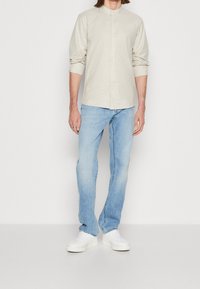 Helles beige Hemd mit Stehkragen, kombiniert mit hellblauen Jeans und weißen Sneakers. Der Stoff wirkt glatt und leicht.