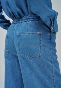 Jean en denim bleu avec une taille élastique, cinq poches et des détails de couture subtils sur les poches arrière. Texture lisse.