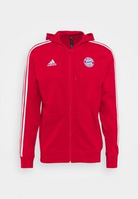 Sweat à zip rouge en tissu doux avec capuche, accents rayés blancs sur les manches et logo du FC Bayern Munich sur la poitrine.