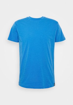 T-shirt blu tinta unita a maniche corte con scollo rotondo, con una piccola etichetta a righe bianca e nera nell'orlo inferiore su sfondo bianco.