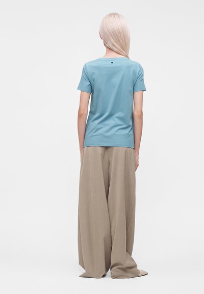 Femme aux longs cheveux blonds portant une chemise bleu clair à manches courtes et un pantalon large beige ample, debout de dos sur un fond blanc.