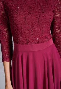 Robe bordeaux avec un corsage en dentelle et des manches trois-quarts, ornée de sequins. La jupe est lisse et plissée, mettant en valeur son mouvement.