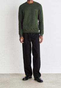 Grüner Wollpullover mit Rundhalsausschnitt und dezentem "BOSS"-Logo auf der Brust, kombiniert mit weiten schwarzen Hosen und schwarzen Schuhen.