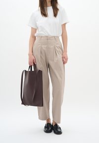 Béžové kalhoty s plisováním, spárované s bílým tričkem. Drží tmavě hnědou koženou tote bag. Černé mokasíny dokončují vzhled.