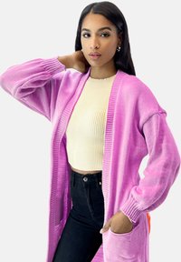 Lange roze cardigan met geribbelde textuur, open voorkant en twee voorzakken, gedragen over een lichtbeige geribbelde top en zwarte high-waisted jeans.