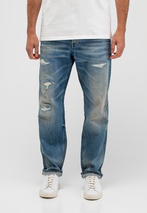 Man die versleten blauwe scheuren jeans, witte sneakers en een wit t-shirt draagt, staand tegen een effen lichtgrijze achtergrond.