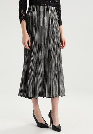 Vrouw draagt een zwarte kanten top, een zilver-zwarte verticale gestreepte geplooide midirok en zwarte hoge hakken, staand op een effen achtergrond.