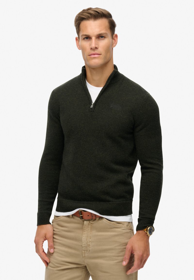 Maglione zip-up verde scuro con colletto alto, polsini e orlo a costine. Indossato sopra una maglietta bianca, abbinato a pantaloni beige chiaro e una cintura marrone.