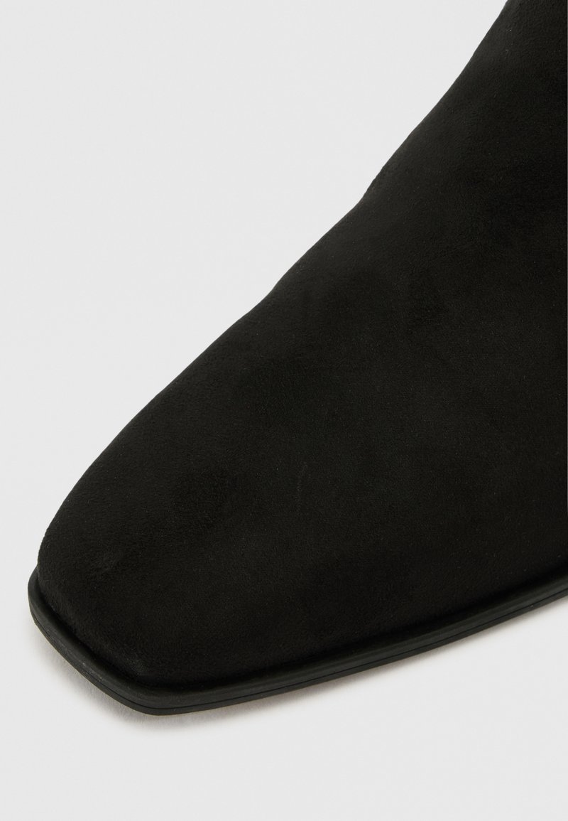 Botín negro de ante hasta el tobillo con punta afilada y acabado suave. Presenta una suela de goma delgada, combinando un diseño moderno con una forma clásica.