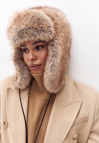 Valkofur-villatakki beige -sävyssä, jossa on paksu ja pehmeä rakenne, leveät korvaläpät ja pyöreä kruunumuotoilu, mikä täydentää ruskean villatakin.