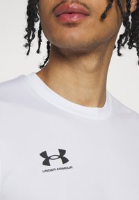 Baltās krāsas īss-iezīmes krekls, izgatavots no gludas auduma, ar melnu Under Armour logotipu uz krūškurvja. Apaļš kakla izgriezums ar plakanām šuvēm.