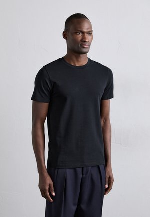 T-shirt noir à manches courtes et col rond en coton, présentant une texture lisse et un design ajusté. Porté avec un pantalon sombre amples.