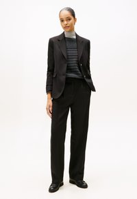 Černý upravený blazer přes šedý pruhovaný rolák, v kombinaci s volnými černými kalhotami a černými botami, vytvářející formální outfit.