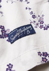 Tela blanca con pequeños patrones florales en púrpura y rojo, y una etiqueta cosida en azul marino que dice "Diseñado y Fabricado por Superdry Manufacturing Co."