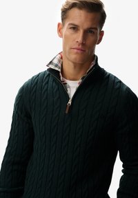 Pull vert foncé en tricot torsadé avec un col demi-fermeture éclair, souligné d'une doublure à carreaux sur le col. Texture douce et poignets côtelés.