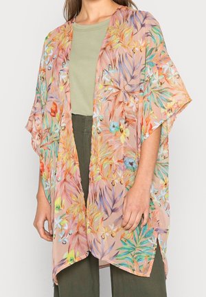 Kvinde iført løs, blomsterprintet, transparent kimono-lignende cardigan over lysegrøn top og mørkegrønne bukser, stående mod en ensfarvet baggrund.
