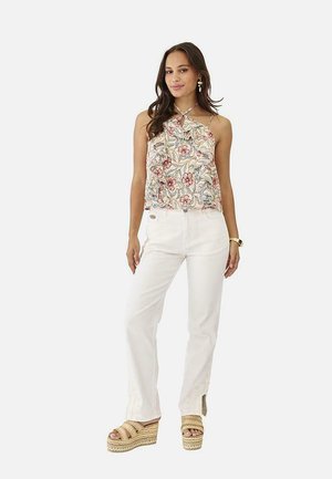 Mujer con top de tirantes floral con volantes, pantalones blancos con aberturas laterales, sandalias de plataforma, pendientes y un reloj de pulsera, de pie sobre un fondo blanco.