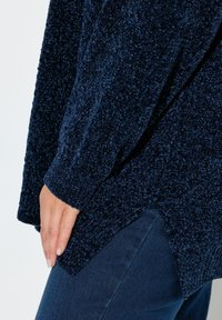 Maglione a maglia testurizzata blu navy con finitura morbida e pelosa, caratterizzato da un orlo asimmetrico e polsini a coste. Indossato sopra un denim scuro.