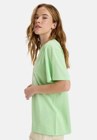 Hellgrünes Baumwoll-T-Shirt mit kurzen Ärmeln, lockerer Passform und Rundhalsausschnitt. Verfügt über ein dezentes grafisches Design auf der Vorderseite.