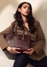 Brown crocodile-embossed leather tote bag with two handles, metal logo accent, structured shape, and smooth texture. Worn with a beige blazer.