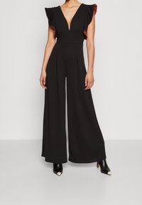 Mujer vestida con un mono negro de pierna ancha con escote en V profundo y mangas de volante, combinado con botas de tobillo negras con punteras blancas.
